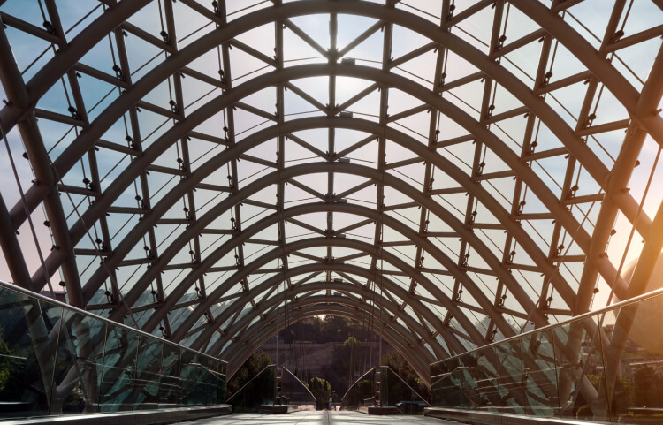 Curved steel beams form a glass ceiling over a bridge at sunset